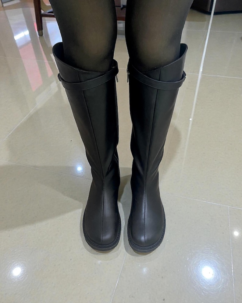 May include: A pair of black knee-high boots featuring a smooth matte finish, a rounded toe, a vertical seam running down the center of the shaft, and a thin horizontal strap detail with a small buckle positioned near the top of the boot shaft.