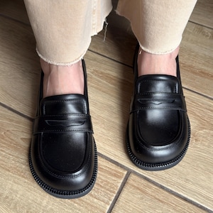 Puede incluir: Un par de mocasines de cuero negro con punta redondeada, una tira cl&aacute;sica tipo penny sobre el empeine y una suela gruesa de goma negra texturizada. Los zapatos tienen un acabado liso y una silueta estructurada con costuras visibles.
