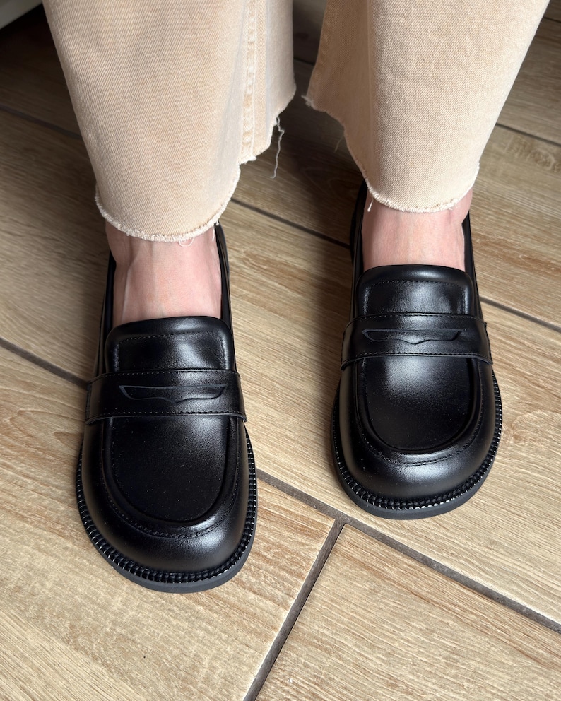 Puede incluir: Un par de mocasines de cuero negro con punta redondeada, una tira cl&aacute;sica tipo penny sobre el empeine y una suela gruesa de goma negra texturizada. Los zapatos tienen un acabado liso y una silueta estructurada con costuras visibles.