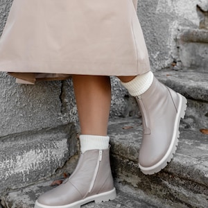 May include: Gray leather ankle boots with white soles and a zipper closure. The boots are worn with white socks and are set against a stone staircase. The boots have a modern, casual style.