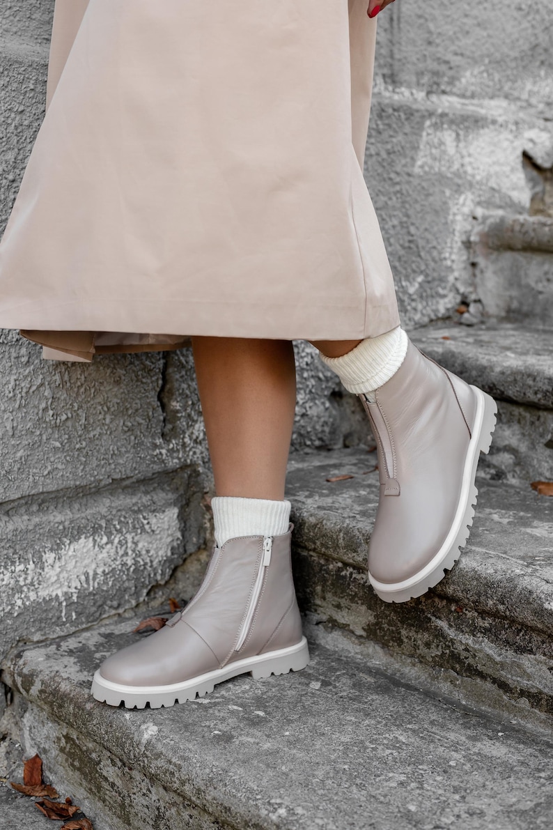 May include: Gray leather ankle boots with white soles and a zipper closure. The boots are worn with white socks and are set against a stone staircase. The boots have a modern, casual style.