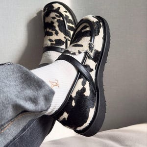 Könnte beinhalten: Ein Paar Loafer mit Kuhfellmuster in Schwarz und Weiß mit einer strukturierten Oberfläche. Die Schuhe verfügen über einen schwarzen Lederriemen über dem Spann, eine dicke schwarze Gummisohle mit Profil und werden mit weißen Socken getragen.