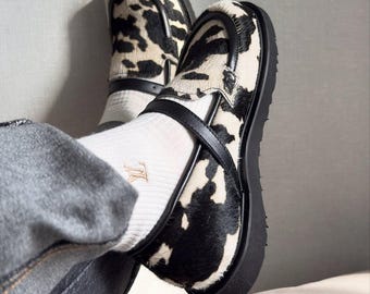 Può includere: Un paio di mocassini con stampa muccata caratterizzati da un motivo a macchie bianche e nere con una finitura testurizzata effetto pelo. Le scarpe includono un cinturino in pelle nera sulla parte superiore, una spessa suola in gomma nera e calze bianche.