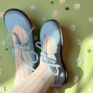 Op de afbeelding: Lichtblauwe ballerina's met bijpassende veters, gecombineerd met cr&egrave;mekleurige panty's met patroon. De schoenen hebben een ronde neus en een zwarte zool. De achtergrond is lichtgroen met verspreide madeliefjesdecoraties.