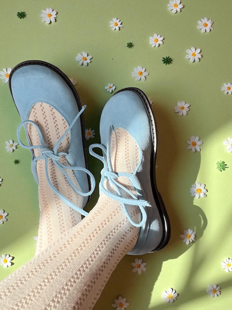 Op de afbeelding: Lichtblauwe ballerina's met bijpassende veters, gecombineerd met cr&egrave;mekleurige panty's met patroon. De schoenen hebben een ronde neus en een zwarte zool. De achtergrond is lichtgroen met verspreide madeliefjesdecoraties.