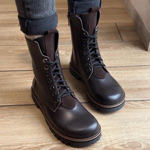 May include: Dark brown leather boots with black laces and suede detailing. The boots feature a round toe, a thick black sole, and a side zip. The boots are worn with dark grey denim jeans. The boots' height is approximately 20 cm.