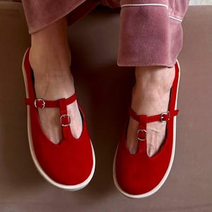 May include: A pair of red Mary Jane style shoes with a T-strap design and silver buckles. The shoes have a rounded toe and a white sole. The shoes are made of a soft material and are suitable for casual wear.
