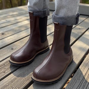 Könnte beinhalten: Braune Chelsea-Stiefel aus Leder mit runder Spitze und schwarzer Sohle. Die Stiefel haben eine Zuglasche oben und ein genähtes Design. Die Stiefel werden mit grauen Jeans getragen.
