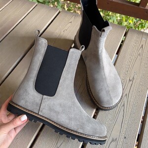 May include: A pair of gray suede Chelsea boots with black elastic side panels and a black rubber sole. The boots have a rounded toe and a pull tab at the back. The boots are displayed on a wooden surface.
