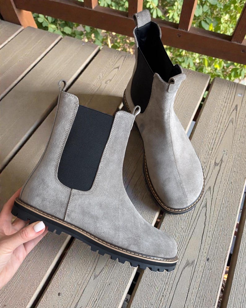 May include: A pair of gray suede Chelsea boots with black elastic side panels and a black rubber sole. The boots have a rounded toe and a pull tab at the back. The boots are displayed on a wooden surface.