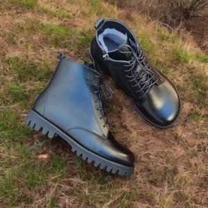 May include: A pair of black leather boots with a lace-up design and a thick, lugged sole. One boot is partially unzipped, revealing a gray interior. The boots are set on a bed of brown and green grass.