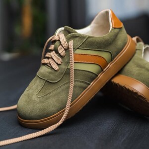 May include: Green suede sneakers with brown and white stripes and brown laces. The sneakers have a brown rubber sole.