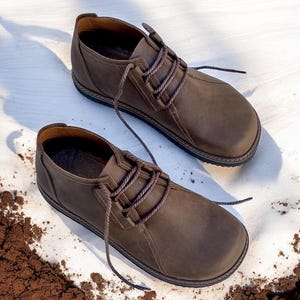 May include: A pair of brown leather shoes with rounded toes and brown laces. The shoes have a stitched design around the sole and are set against a white background with a patch of brown soil.