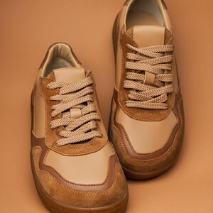 May include: A pair of tan and brown sneakers with matching laces. The shoes have a suede and leather design, with a brown rubber sole. The sneakers are displayed on a solid brown background.