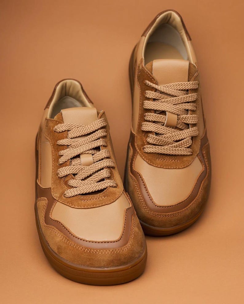 May include: A pair of tan and brown sneakers with matching laces. The shoes have a suede and leather design, with a brown rubber sole. The sneakers are displayed on a solid brown background.