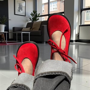 Op de afbeelding: Felrode Mary Jane schoenen met een ronde neus en een striksluiting. De schoenen hebben een zwarte zool en worden gecombineerd met een donkere jeans. Een klassieke stijl.