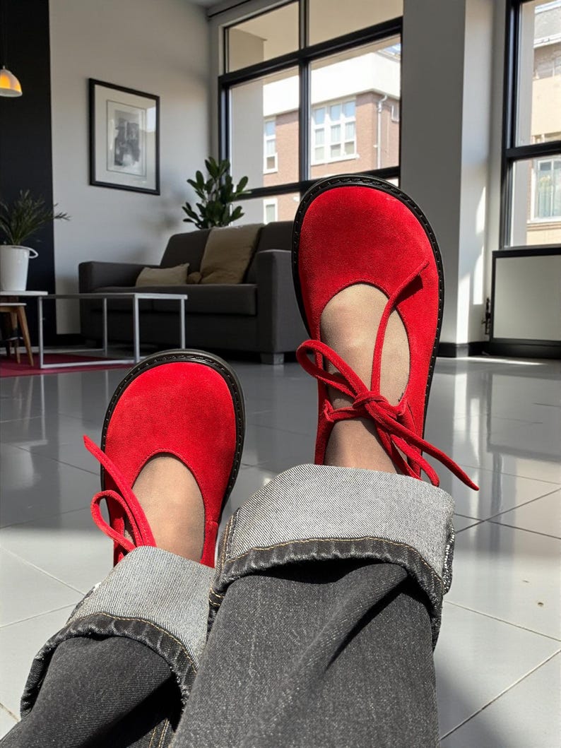 Op de afbeelding: Felrode Mary Jane schoenen met een ronde neus en een striksluiting. De schoenen hebben een zwarte zool en worden gecombineerd met een donkere jeans. Een klassieke stijl.
