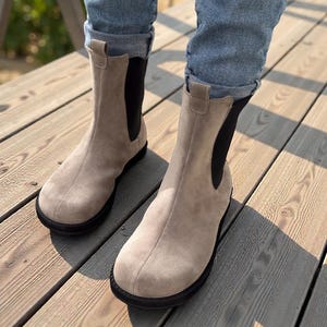 May include: A pair of beige suede Chelsea boots with black elastic side panels and black soles. The boots are worn with blue jeans, and are on a wooden deck.