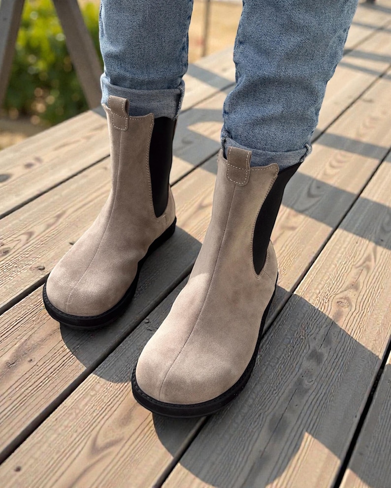 May include: A pair of beige suede Chelsea boots with black elastic side panels and black soles. The boots are worn with blue jeans, and are on a wooden deck.