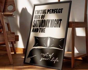 May include: A black framed art print with the text "I'm the perfect mix of Saturday Night and the rest of your life." The print features a black and white illustration of a bed with pillows. The print is leaning against a wall.