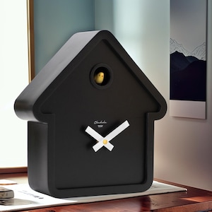 May include: A black cuckoo clock in the shape of a house. The clock has white hands and a yellow center. A small yellow bird is visible in a round opening. The clock has the word "Cluckola" written on the front.