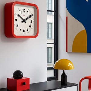 May include: A red square wall clock with a white face and black hands and numbers. A black sphere sits atop a red cube with a black knob. A yellow lamp with a black base is on a black and white cabinet.