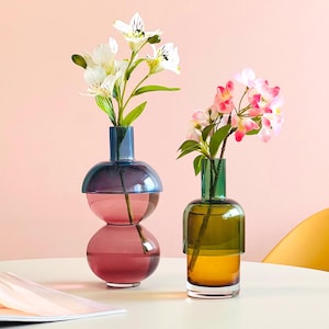 May include: Two glass vases with flowers. The vase on the left is a double-bubble design with a teal top and a pink bottom. The vase on the right is a tall, cylindrical design with a green top and a brown bottom. Both vases are filled with flowers.