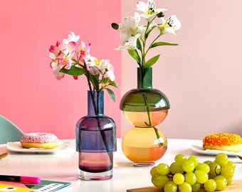 May include: Two glass vases with flowers on a white table. The vase on the left is blue and purple with pink flowers. The vase on the right is green and orange with white flowers. There are grapes on a wooden cutting board in the foreground.