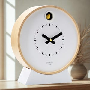 May include: A circular white clock with a light wood frame and a white base. The clock face features black hands and a small yellow bird. The clock is on a wooden surface. The clock has the text "Claudia" on the face.