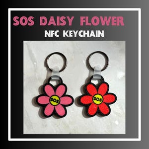 May include: Two daisy flower keychains with black metal rings and clear plastic loops. The flowers are pink and red with black outlines and a yellow center with the word "SOS" in black letters.