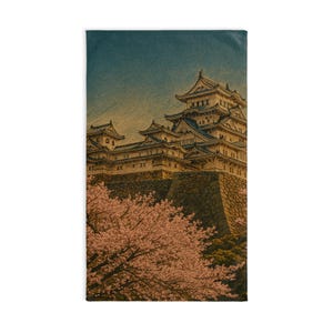 以下が含まれることがあります： 日本の城のイラストが描かれた長方形のタオル。青い屋根の多層構造の城が、グラデーションの空を背景に描かれています。ピンク色の桜と緑の木々が前景にあります。