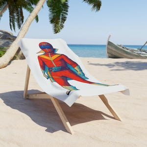 Puede incluir: Una toalla de playa blanca con una colorida ilustración de un superhéroe con un traje rojo y azul. La toalla está sobre una silla de playa de madera en una playa de arena, con un barco al fondo.
