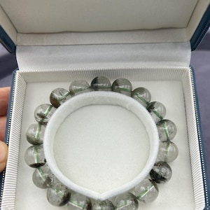 Op de afbeelding: Een heldere kwarts armband met groene insluitsels in een blauwe fluwelen doos. De armband is gemaakt van ronde kralen en heeft een eenvoudig ontwerp.