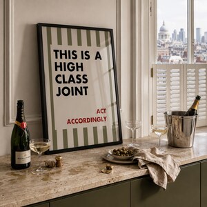 May include: A framed rectangular wall art print featuring vertical olive green stripes and the black text THIS IS A HIGH CLASS JOINT with the red text ACT ACCORDINGLY below. The print is displayed in a thin black frame with a clean, modern aesthetic.