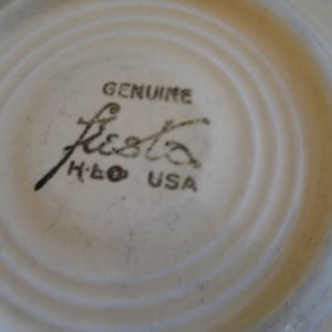 May include: Close-up of a cream-colored Fiesta saucer with concentric rings. The center features the words "GENUINE Fiesta H.L.C. USA" in black script and block letters, indicating its authenticity.