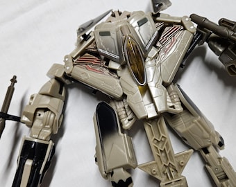 May include: A detailed image of a beige and gray action figure with articulated joints. The figure has a complex design with visible mechanical details, including red and silver accents. It is posed with arms extended, showcasing its intricate construction.