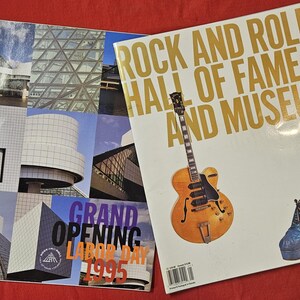 May include: Two magazines on a red surface. One features architectural images and text that reads "Grand Opening Labor Day 1995". The other has "Rock and Roll Hall of Fame and Museum" with a guitar and shoes.