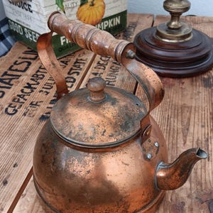 May include: A vintage copper kettle with a wooden handle and spout. The kettle has a rounded body and a lid with a small knob. The copper has a weathered patina, suggesting age. The kettle is sitting on a wooden surface.