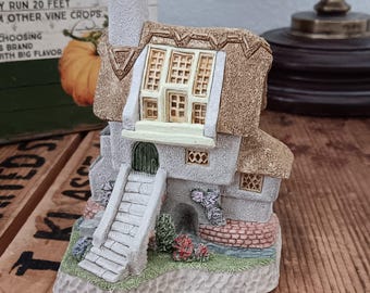 May include: A miniature cottage figurine with a thatched roof, stone walls, and a chimney. The cottage has a small door, windows, and a staircase leading to the entrance. The base is decorated with greenery and flowers. The figurine is set on a wooden surface.