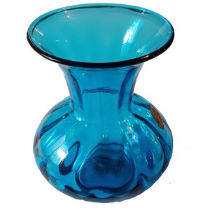 May include: A turquoise glass vase with a wide, open top and a rounded body. The vase has a dark rim and a textured surface, reflecting light. A small, round sticker is visible on the side. The vase is likely used for decorative purposes.