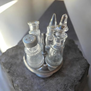 May include: A clear glass cruet set featuring four small bottles held in a circular metal stand. One bottle has a silver-toned perforated cap, while the others have clear glass stoppers. The set is arranged with a central handle for carrying.