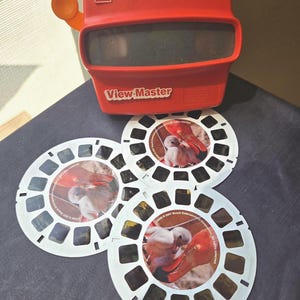 May include: A red GAF View-Master stereoscope viewer with an orange advance lever, accompanied by three circular white reels featuring photographic images of a flamingo and its chick, marked with 2007 Busch Entertainment Corporation copyright text.