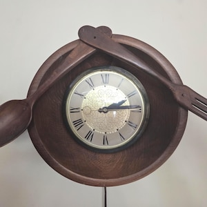 May include: A wall clock featuring a circular dark wood bowl as the frame, with a large wooden spoon and fork crossed at the top. The clock face has a gold-toned centre with intricate filigree patterns and black Roman numerals for the hour markers.