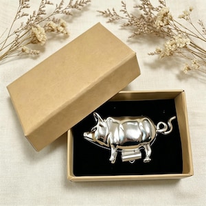 May include: A silver-colored pig-shaped money clip presented in a brown cardboard gift box with a black interior. The pig has a curled tail and is standing on four legs. The box lid is open.