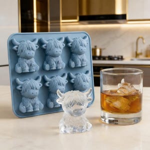 Puede incluir: Una cubitera de silicona azul claro con seis formas de vaca de las Tierras Altas. Un cubo de hielo transparente con forma de vaca está al lado de la cubitera, y un vaso de líquido ámbar con un cubito de hielo con forma de vaca está a la derecha.