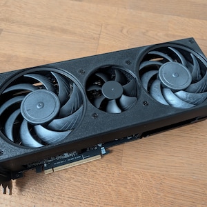 Könnte beinhalten: Eine schwarze Grafikkarte mit drei Kühlventilatoren. Die Ventilatoren sind in einer Reihe angeordnet und haben gebogene Flügel. Die Karte hat eine rechteckige Form und ist auf einer Leiterplatte montiert.