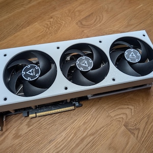 Op de afbeelding: Een zilver-zwarte grafische kaart met drie grote ventilatoren. Elke ventilator heeft zwarte bladen en een centraal logo. De kaart is ontworpen voor computergebruik en heeft een rechthoekige vorm met een metalen behuizing.