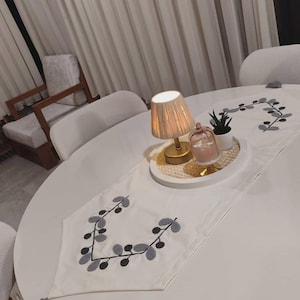 May include: A white table runner with embroidered gray and black leaf design. A small gold lamp, a candle in a glass cloche, and a small potted plant sit on a round tray. The table is white, and the background includes chairs and curtains.