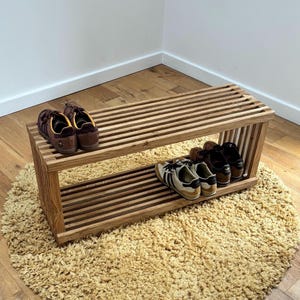 Puede incluir: Un zapatero de madera con dos estantes. El estante superior tiene dos pares de zapatos marrones y el estante inferior tiene dos pares de zapatos negros y blancos. El zapatero está sobre una alfombra marrón claro.