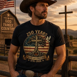 May include: A black t-shirt with a patriotic design featuring a cross, tree, and American flag elements. The shirt includes the text "250 YEARS FAITH AND FREEDOM 1776-2026". A man wearing a cowboy hat and jeans is shown.